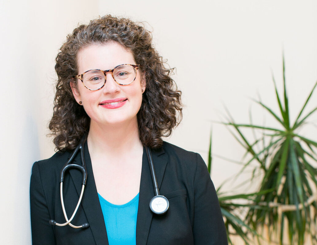 Dr. Barrett, a white female with brown curly hair, smiling. She wears dark, heavy glasses and a turquoise shirt with a black blazer.